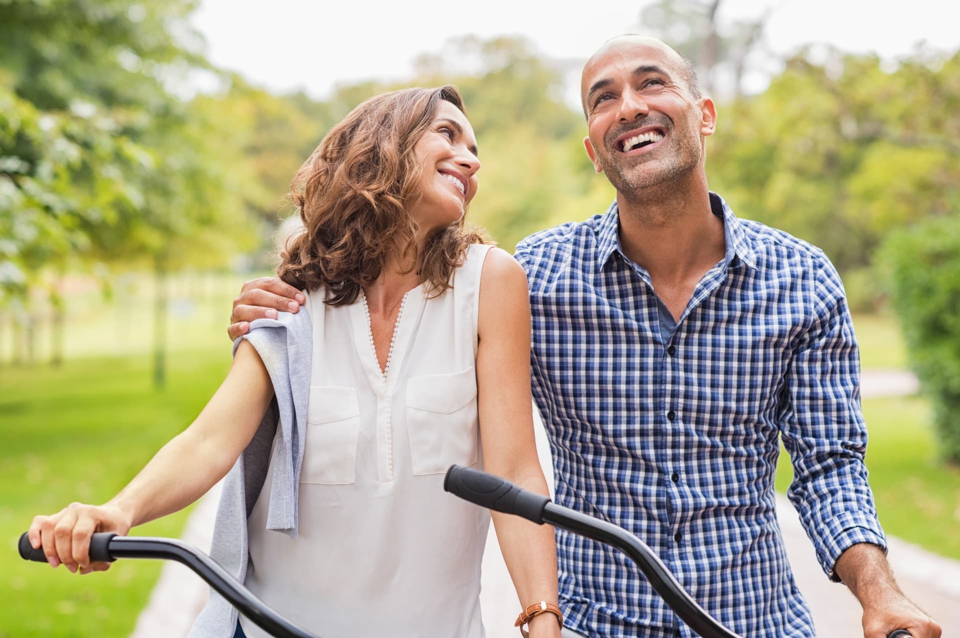Smiling middle-aged couple walking with bicycles through a tree-lined park on a sunny day at Livana Fair Lawn in New Jersey.